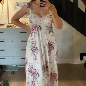 Vit blommig långklänning - Superfin långklänning i vitt med blommor. Klänningen har volangaxelband, spetsdetaljer vid urringningen och en lätt slits framtill. Aldrig använd, tyvärr lite för stor för mig.