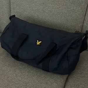 Mörkblå träningsväska Lyle & Scott - Säljer en mörkblå träningsväska från Lyle & Scott med klassisk gul logga framtill och tydlig varumärkespatch på andra sidan. Väskan har både handtag och axelrem, dragkedja upptill och är tillverkad i slitstark polyester. Perfekt storlek för gymmet eller sportaktiviteter.