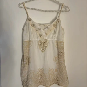 Vintage topp med paljetter  - Supersöt linnetopp i vitt och beige med tunna axelband. Toppen har v-ringning, broderade detaljer och paljetter framtill samt ett unikt mönster med hjärtan och blommor. Lätt och luftig, perfekt för varma dagar eller när du vill sticka ut lite extra.