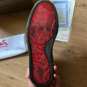 Grå mocka sneakers från Louboutin - Snygga grå sneakers i mocka från Christian Louboutin med klassisk röd sula och svarta snören. Skorna har rund tå och platt sula, samt diskreta metalldetaljer vid snörningen. Perfekta för dig som vill sticka ut med exklusiv streetstyle.
