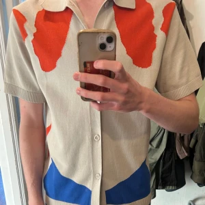 Limitato kortärmad skjorta beige/röd/blå - Unik kortärmad skjorta från Limitato med konstnärligt mönster i rött och blått på beige botten. Skjortan har klassisk krage, knäppning framtill och ribbade ärmslut. Materialet är stickat och känns mjukt mot huden. Limited edition från samarbetet med Loribelle Spirovski. Aldrig används ute så skicket är som nytt! Inga låda till plagget finns! Köpt för 4400kr på NK