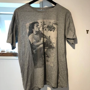 Grå t-shirt med retro fototryck - Grå t-shirt i storlek L med ett stort svartvitt fototryck framtill som visar en person sittande med sportiga detaljer. T-shirten har rund halsringning och korta ärmar. Materialet känns mjukt och lätt, perfekt för en avslappnad stil.