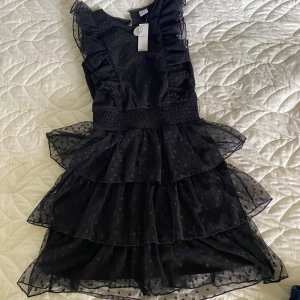 ny Svart ruffle dress med prickar  - Supersöt svart kortklänning från Lindex med volanger och små prickar i tyget. Klänningen har ärmlös design, markerad midja med smock och flera lager volanger i kjolen för extra volym. barnstorlek 152, men passar bra xs