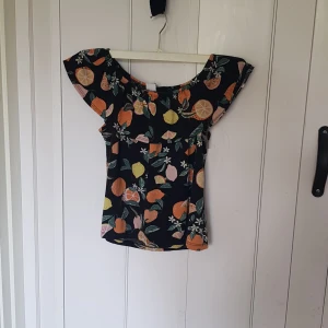 Blommig topp med citrusmönster Lindex - Snygg topp från Lindex med färgglatt citrusmönster i orange, gul och rosa på svart botten. Modellen har breda volangärmar och en offshoulder-look. Perfekt för dig som gillar färg och vill sticka ut lite extra i sommarvärmen!