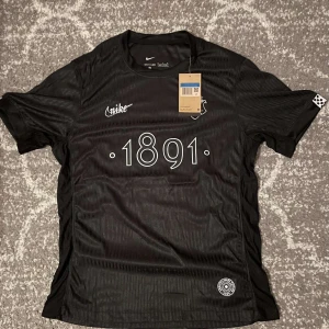 Svart AIK fotbollströja Nike 1891 - AIK fotbollströja från Nike i svart med diskreta ränder och trycket '1891' på bröstet. Tröjan har klubbmärket på vänster sida och Nike-logga på höger. Tillverkad i lätt och ventilerande material, perfekt för match eller träning.
