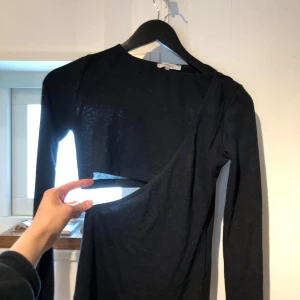 Svart långärmad topp med cut out Chiquelle - Cool svart långärmad topp från Chiquelle med unik asymmetrisk cut out-detalj över magen. Tunn och stretchig, perfekt för dig som vill sticka ut. Snygg passform och enkel att matcha med både jeans och kjol.