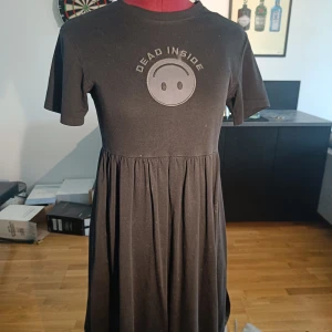 Svart kortklänning med tryck - Svart kortklänning med rund hals och korta ärmar. Framtill finns ett tryck med texten 'DEAD INSIDE' och en smiley. Klänningen har en rynkad midja som ger en avslappnad siluett. Perfekt för dig som gillar en edgy och bekväm stil.