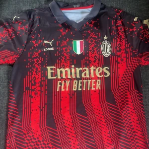 AC Milan x Puma matchtröja röd/svart - AC Milan fotbollströja från Puma i svart och rött med digitalt pixelmönster. Guldiga tryck med Emirates och klubbmärke framtill, samt sponsorloggor på ärmarna. Tillverkad i lätt och ventilerande polyester, perfekt för match eller träning. Det är storlek S men den passar säkert för en storlek M