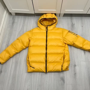 Gul pufferjacka från Emporio Armani - Säljer en gul pufferjacka från Emporio Armani EA7 med huva och svarta detaljer. Jackan har dragkedja framtill, två fickor med dragkedja och logga på ärmen. Perfekt för kalla dagar när du vill sticka ut med en färgstark look. Endast använd 2 gånger och passar även XXL. Nypris 4499kr