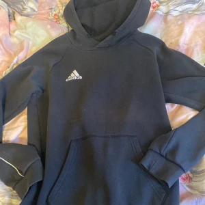 Svart Adidas hoodie med broderad logga - Svart hoodie från Adidas med broderad vit logga på bröstet. Klassisk känguruficka framtill, huva med snörning och mjukt fleecefoder på insidan. Långa ärmar med diskret vit detaljsöm. Perfekt för chill eller träning.