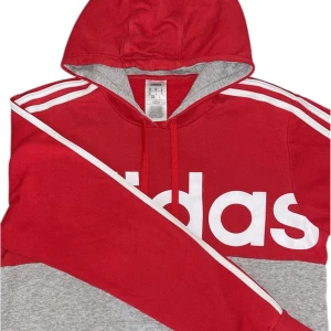 Röd och grå Adidas hoodie med huva - Snygge röd och grå hoodie från Adidas med stor vit logga framtill och klassiska vita ränder på axlarna. Tröjan har huva med dragsko och är tillverkad i mjuk bomullsmix. Perfekt för en sportig och avslappnad stil.