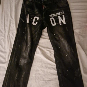 Svarta Dsquared2 ICON jeans med dragkedja - Snygga svarta jeans från Dsquared2 med stor vit 'ICON'-text framtill och coola färgstänk. Jeansen har en unik dragkedjedetalj med ring på fickan och patch med Dsquared2-logga bak. Raka ben och klassisk femficksmodell, perfekt för dig som gillar streetstyle.