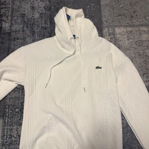 Vit stickad hoodie från Lacoste - Vit stickad hoodie från Lacoste med klassiskt kabelstickat mönster och broderad krokodil på bröstet. Tröjan har huva med snörning och långa ärmar. Perfekt för dig som gillar stilren och sportig look. Knappast använd liten i storleken 