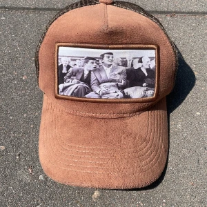 Brun truckerkeps från Maggiore - Snygg brun truckerkeps från Maggiore med mockaliknande material framtill och mesh baktill. Framsidan har en stor svartvit fotoprint med vintagekänsla. Justerbar snapback för perfekt passform. Perfekt accessoar för en chill streetstyle. Spännet på baksidan har spruckit lite grann men funkar bra! Kepsen kommer med en maggiore låda.