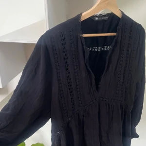 Svart klänning med spets från Zara - Svart klänning från Zara med v-ringning och dekorativa spetsdetaljer längs halsringning, ärmslut och framtill. Långärmad och luftig modell i bomull, broderade partier och går ner till lite över knäna.