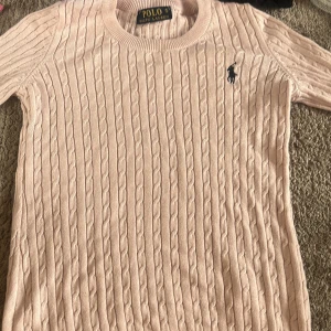 Ljusrosa stickad tröja Polo Ralph Lauren - Supersöt ljusrosa stickad tröja från Polo Ralph Lauren med klassiskt kabelstickat mönster och broderad marinblå logga på bröstet. Tröjan har rund halsringning och långa ärmar, perfekt för lager-på-lager under hösten och våren.