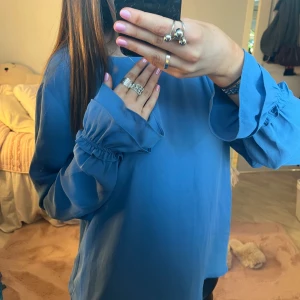 Blå blus med volangärm - Supersnygg blå blus från Gina Tricot i storlek 40. Blusen har långa ärmar med volangdetalj vid ärmslut och en liten öppning i nacken. Luftigt material som ger en skön och flowy känsla. Perfekt att styla med skinnbyxor eller jeans.