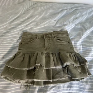 Olivgrön volangkjol från H&M Divided - Säljer en olivgrön jeanskjol från H&M Divided med dubbla volanger och råa kanter. Kjolen har klassiska jeansfickor fram och bak samt knapp och dragkedja i midjan. Perfekt för dig som gillar en edgy och trendig stil.