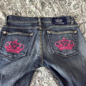 Victoria beckham jeans rosa  - Mörkblå jeansbyxor från Victoria Beckham med raka ben och snygga slitningar. Bakfickorna har broderade rosa kronor med paljettdetaljer. Klassisk femficksmodell med knappar och dragkedja framtill.