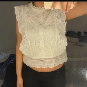 Vit spetsblus med volangdetaljer - Säljer en söt vit blus i spets med volangdetaljer längs ärmhål och nederkant. Blusen är ärmlös och har ett vackert spetsmönster över hela plagget. Perfekt för vår och sommar, och ger en romantisk känsla till outfiten.
