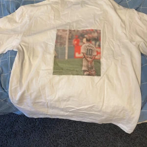 Vit t-shirt med Zidane tryck på ryggen - Vit t-shirt med en stor tryckt bild på ryggen av en fotbollsspelare med nummer 10 och namnet Zidane. Klassisk passform och rund halsringning. Perfekt för dig som gillar fotboll och vill ha en unik t-shirt. 