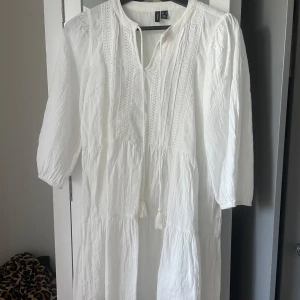 Vit klänning med spetsdetaljer  - Säljer en luftig vit klänning med fina spetsdetaljer framtill och knytband vid halsen. Klänningen har trekvartsärm och en avslappnad passform, perfekt för varma dagar.