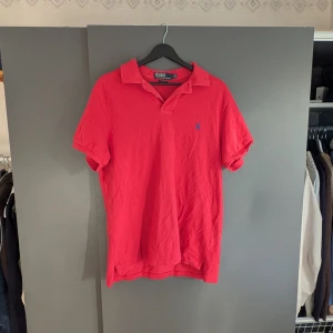 Röd pikétröja från Polo Ralph Lauren - Säljer en klassisk röd pikétröja från Polo Ralph Lauren i slim fit-modell. Tröjan har korta ärmar, krage och knappslå samt den ikoniska broderade loggan på bröstet. Perfekt för en stilren och sportig look.