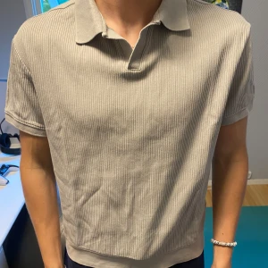 Beige ribbad pikétröja från Zara - Säljer en stilren beige pikétröja från Zara med ribbad struktur. Tröjan har korta ärmar och klassisk krage med knappslå. Perfekt för en avslappnad men snygg look.