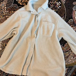 Beige teddyjacka med ficka - Mysig beige teddyjacka med klassisk krage och tryckknappar framtill. Jackan har en bröstficka och är långärmad. Perfekt för kyliga dagar när du vill vara både varm och snygg.