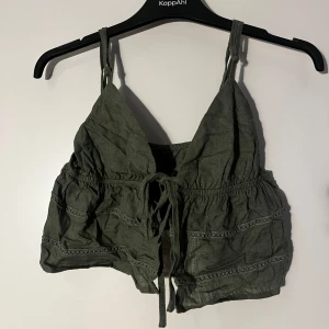 Olivgrön croppad linnetopp med knyt - Säljer en olivgrön croppad linnetopp med smala axelband och knyt framtill. Perfekt för varma dagar och enkel att styla till olika outfits. 