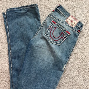 True Religion Becca Mid Rise Bootcut Jeans - Säljer ett par blå True Religion Becca jeans med bootcut och mellanhög midja. Jeansen har röda sömmar och detaljerade bakfickor med logga. Storlek 29, jag är 164cm. Använda några få gånger så väldigt bra skick. Säljer då dom ej kommer till användning. 