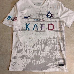 Al Nassr fotbollströja - Säljer en vit bortatröja från Al Nassr med grått mönster, Nike-logga och klubbmärke på bröstet. Tryck med KAFD och Mane 10 på ryggen. Slim fit-modell i lätt och ventilerande material.