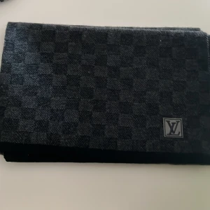 Lv halsduk - Snygg mörkgrå halsduk från Louis Vuitton med diskret rutigt mönster och klassisk LV-logga. Halsduken är tillverkad i ull och har en exklusiv känsla, perfekt för att lyfta din outfit.