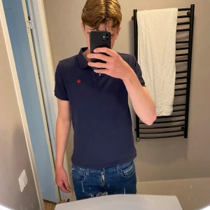 Mörkblå pikétröja morris - Säljer en mörkblå pikétröja med klassisk krage och två knappar framtill. Tröjan har en liten röd broderad logga på bröstet och är kortärmad. Perfekt för en stilren och avslappnad look. Det är märket Morris som är populärt och eftertraktat märke som är lite av dem dyrare och finare märkena.