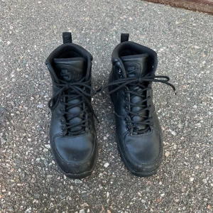 Svarta boots från Nike - Säljer ett par svarta boots från Nike med snörning och rund tå. Skorna är helt svarta med diskret Nike-logga på sidan och grov sula för extra grepp. Perfekta för höst och vinter när du vill ha både stil och funktion.
