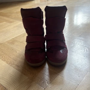 Vinröda sneakers med klack - Säljer ett par vinröda sneakers med klack och kardborreband. Skorna har en blandning av mocka och syntetmaterial, samt en platt sula framtill. Perfekta för dig som vill ha både stil och komfort.