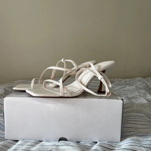 Vita högklackade sandaler med smala remmar - Säljer ett par vita sandaler med smala remmar och öppen tå. Skorna har en elegant klack och justerbart spänne vid ankeln. Perfekta för dig som vill ha en stilren och trendig look. Nyskick, aldrig använda och endast testade🫶