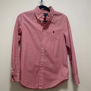 Röd- och vitrandig skjorta från Ralph Lauren - Klassisk röd- och vitrandig skjorta från Ralph Lauren med lång ärm och knappar framtill. Skjortan har en broderad logga på bröstet och är i en custom fit-modell. Perfekt för dig som gillar stilrena och tidlösa plagg. Storlek 12 år.