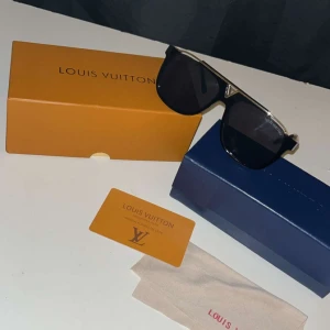 Svarta solglasögon från Lv - Snygga svarta solglasögon från Lv med rektangulära glas och en tydlig gulddetalj på ovansidan. Bågarna är svarta med en touch av guld, och glasen är mörka. Perfekt accessoar för att lyfta din stil.