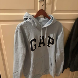 Ljusblå hoodie från GAP - Säljer en ljusblå hoodie från GAP i storlek M. Tröjan har klassisk huva med dragsko, stor GAP-logga framtill och känguruficka. Tillverkad i mjuk bomullsblandning för extra komfort. Perfekt för en avslappnad stil.
