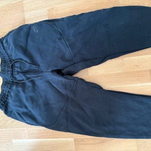 Svarta Nike tech mjukisbyxor - Svarta mjukisbyxor från Nike med dragsko i midjan och diskret Nike-logga på vänster sida. Byxorna har en avslappnad passform och är tillverkade i mjukt bomullsmaterial, perfekta för chill eller träning.