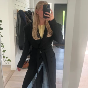 Svart kappa med bälte - Säljer en svart lång kappa med bälte i midjan. Breds axlar och smal midja med långa ärmar och krage. Perfekt att slänga på sig över jeans eller klänning för en snygg look. Köpt för 3000kr