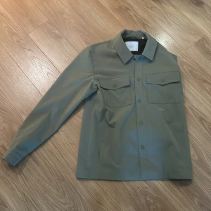 Grön overshirt från Les Deux - Stilren grön overshirt från Les Deux med klassisk krage och två stora bröstfickor med lock. Jackan har knäppning framtill och lång ärm. Perfekt för lager-på-lager och enkel att matcha med olika outfits. Pris kan diskuteras vid snabb affär!!!
