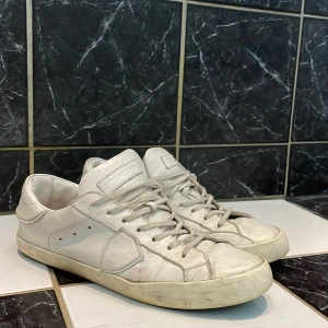 Vita sneakers med snörning - Säljer ett par klassiska vita sneakers från Philippe model med snörning och rund tå. Skorna har en platt sula och är tillverkade i skinn. Perfekta för en stilren och enkel look.