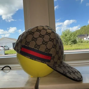 Gucci keps med GG-mönster och röd-blå rand - Snygg keps från Gucci med klassiskt GG-mönster i beige. Kepsen har en röd och blå rand på sidan och är tillverkad i ett slitstarkt tyg. Perfekt accessoar för att lyfta din stil.