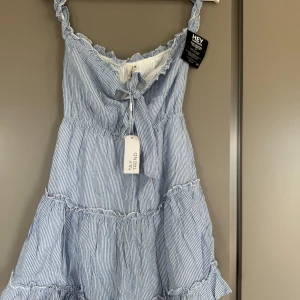 Blåvit randig klänning från NLY Trend - Säljer en söt blåvit randig klänning från NLY Trend med volangdetaljer och knyt framtill. Klänningen har smala axelband och kort, perfekt för varma dagar.