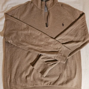 Beige halvzip tröja från Polo Ralph Lauren - Säljer en beige långärmad halvzip tröja från Polo Ralph Lauren med klassisk broderad logga på bröstet. Tröjan har hög krage och muddar vid ärmslut och nederkant. Perfekt för lager-på-lager och stilren vardagslook. Storlek M