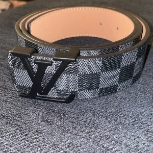 Svart rutigt bälte från Louis Vuitton - Säljer ett svart bälte från Louis Vuitton med klassiskt rutigt mönster och stor LV-logga på spännet. Bältet har en stilren design och insidan är ljusbrun. Perfekt accessoar för att lyfta din outfit.