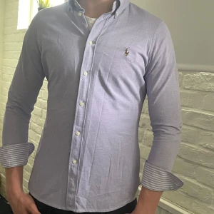 Ljuslila skjorta från Ralph Lauren - Ralph Lauren Oxfordskjorta – Slim Fit, Ljuslila Storlek på skjortan: Är L Modellens längd: 187 cm  Skick: 9/10 Inga fläckar eller hål Material: 100 % bomull Färg: Ljuslila med vit- och lilamönstrad insida på ärmslut Passform: Slim fit, sitter nära kroppen Detaljer: – Klassisk button-down-krage – Broderad Polo-logga på bröstet