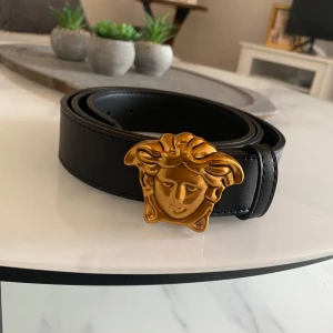  Versace bälte - Säljer ett svart bälte från Versace med ett stort guldigt Medusa-huvud som spänne. Bältet är gjort i skinn och har en stilren design som sticker ut tack vare det ikoniska spännet. Perfekt accessoar för att lyfta din outfit.